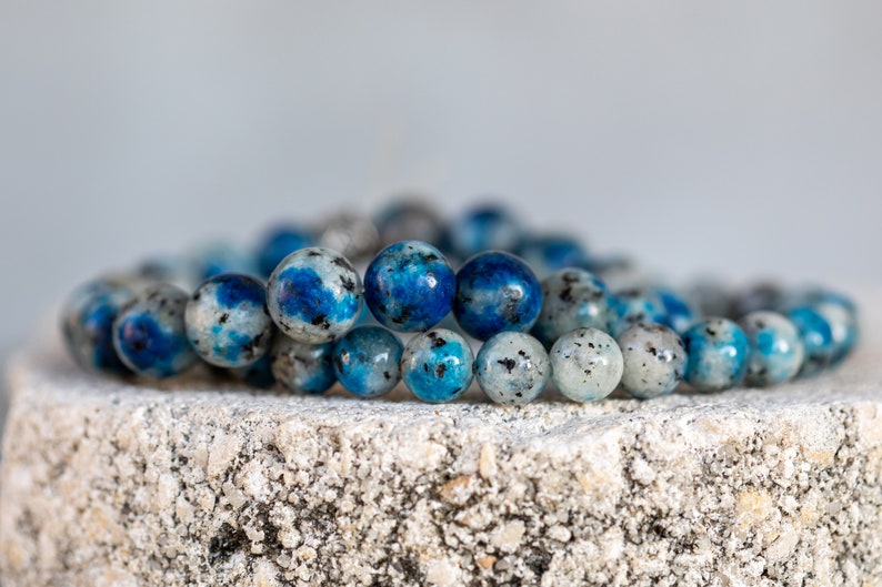 May include: A beaded bracelet made of blue and white kyanite stones. The bracelet is made of round beads and is stacked on top of another bracelet.