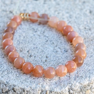 May include: A beaded bracelet featuring peach-colored, round gemstone beads. The bracelet is strung on a clear elastic cord and has gold-colored metal accents. The bracelet is resting on a textured, light gray surface.