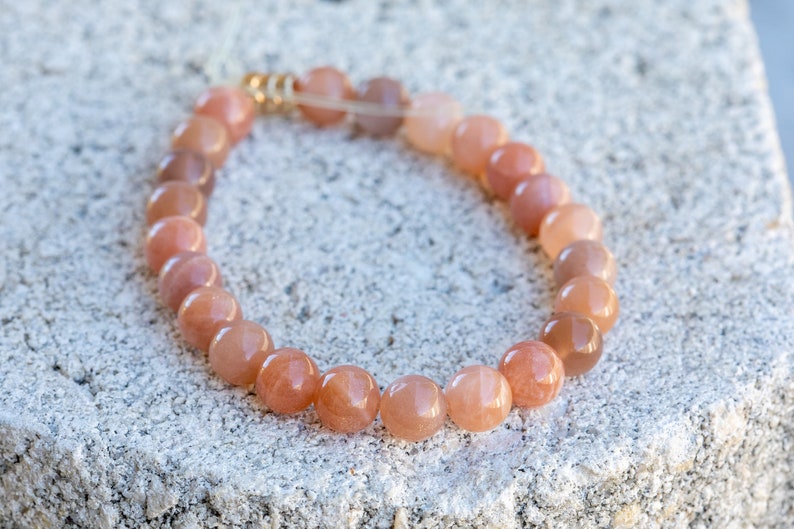 May include: A beaded bracelet featuring peach-colored, round gemstone beads. The bracelet is strung on a clear elastic cord and has gold-colored metal accents. The bracelet is resting on a textured, light gray surface.
