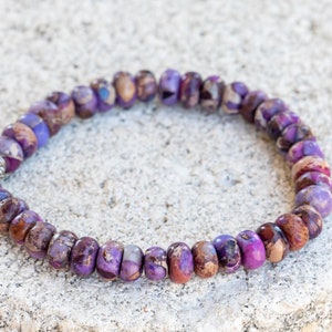 May include: A purple and brown gemstone bracelet with a silver bead accent. The bracelet is made of small, round beads and is strung on a thin cord.