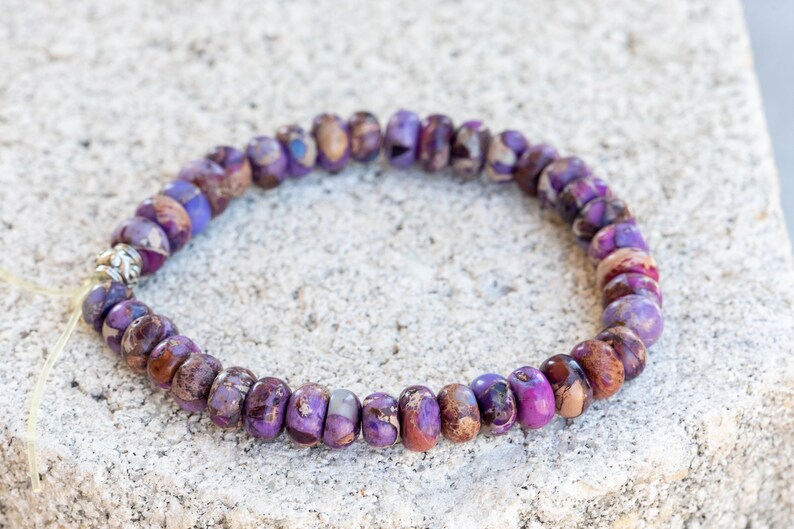 May include: A purple and brown gemstone bracelet with a silver bead accent. The bracelet is made of small, round beads and is strung on a thin cord.