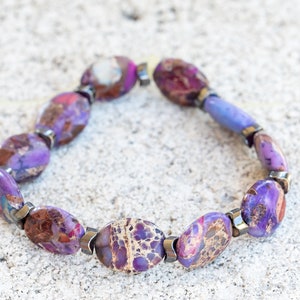 May include: A purple and brown gemstone bracelet with silver accents. The bracelet is made of oval-shaped stones and is strung on a silver chain.