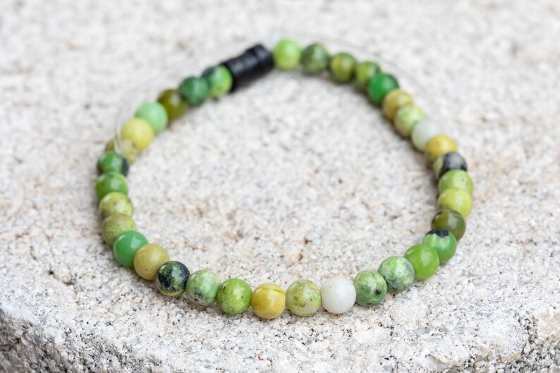 K&ouml;nnte beinhalten: Ein gr&uuml;nes Edelstein-Armband mit einem Farbverlauf von Dunkelgr&uuml;n &uuml;ber Hellgr&uuml;n bis Gelb. Das Armband besteht aus runden Perlen und hat einen schwarzen Verschluss.