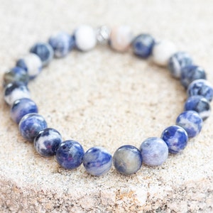 May include: A blue and white gemstone bracelet with a silver clasp. The bracelet is made of round beads and is lying on a light brown surface.