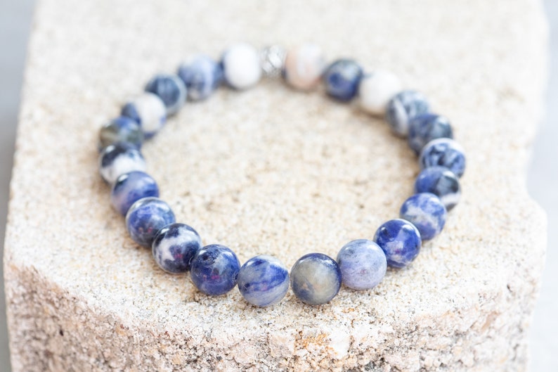 May include: A blue and white gemstone bracelet with a silver clasp. The bracelet is made of round beads and is lying on a light brown surface.
