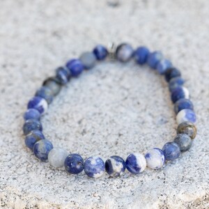 May include: A blue and white gemstone bracelet with a silver clasp. The bracelet is made of round beads and is approximately 7 inches in length.