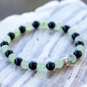 May include: A bracelet made of black and green beads with a silver charm. The bracelet is on a white marble surface.