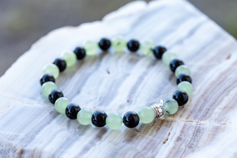 May include: A bracelet made of black and green beads with a silver charm. The bracelet is on a white marble surface.