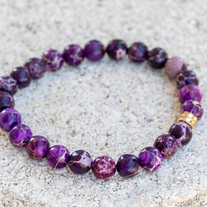 May include: Purple and white patterned gemstone bracelet with a gold accent bead.