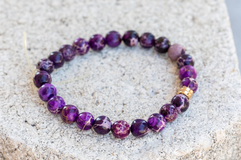 May include: Purple and white patterned gemstone bracelet with a gold accent bead.