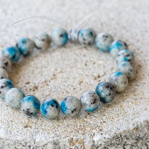 May include: A blue and white gemstone bracelet with a silver clasp. The bracelet is made of round beads and is lying on a rough, textured surface.