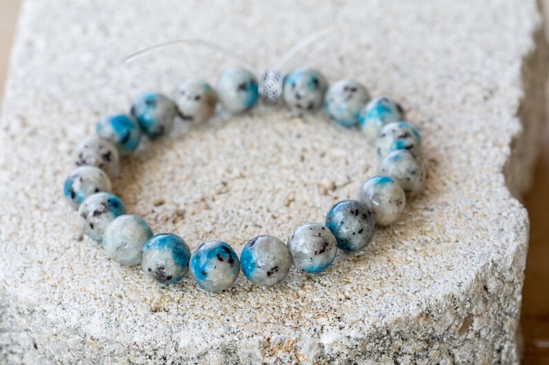 May include: A blue and white gemstone bracelet with a silver clasp. The bracelet is made of round beads and is lying on a rough, textured surface.