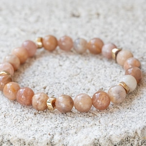 May include: A beaded bracelet featuring peach-colored, round gemstones interspersed with small gold-colored metal accents. The bracelet is displayed on a textured, light-colored surface.