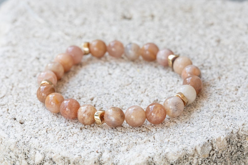 May include: A beaded bracelet featuring peach-colored, round gemstones interspersed with small gold-colored metal accents. The bracelet is displayed on a textured, light-colored surface.