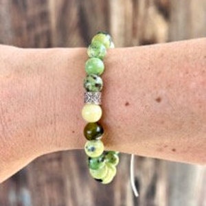 K&ouml;nnte beinhalten: Ein gr&uuml;nes und wei&szlig;es Edelstein-Armband mit einem silbernen Zwischenst&uuml;ck. Das Armband besteht aus runden Perlen und ist auf einer wei&szlig;en Schnur aufgef&auml;delt.