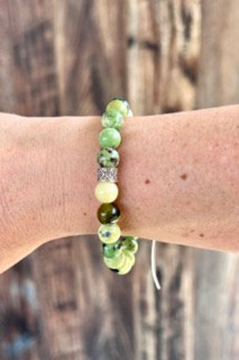 K&ouml;nnte beinhalten: Ein gr&uuml;nes und wei&szlig;es Edelstein-Armband mit einem silbernen Zwischenst&uuml;ck. Das Armband besteht aus runden Perlen und ist auf einer wei&szlig;en Schnur aufgef&auml;delt.