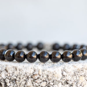 May include: A black beaded bracelet made of round, smooth stones. The bracelet is lying on a rough, gray surface.