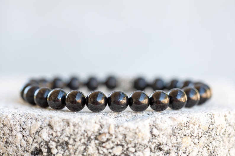 May include: A black beaded bracelet made of round, smooth stones. The bracelet is lying on a rough, gray surface.