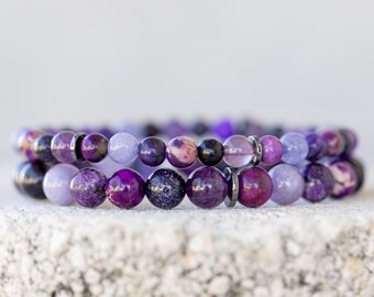 May include: Two stacked bracelets featuring a variety of purple beads. The beads range in color from deep violet to lavender, with some having a speckled or iridescent appearance. The bracelets are resting on a textured, light-colored surface.