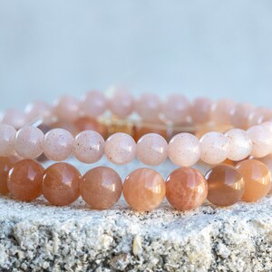May include: Two beaded bracelets with round, polished beads. The top bracelet features light pink beads, while the bottom bracelet has darker, peach-colored beads. The bracelets are stacked on a textured, light-colored surface.