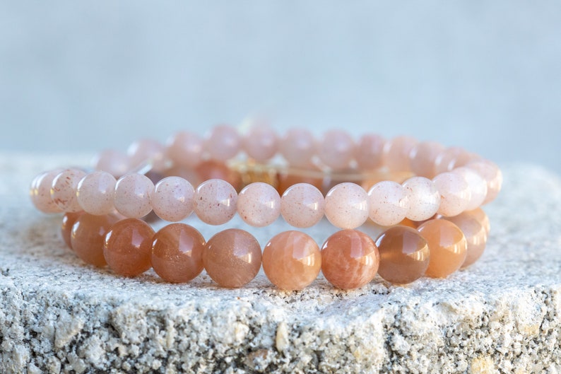 May include: Two beaded bracelets with round, polished beads. The top bracelet features light pink beads, while the bottom bracelet has darker, peach-colored beads. The bracelets are stacked on a textured, light-colored surface.
