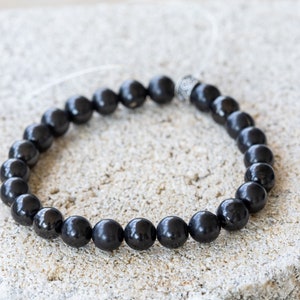 May include: A black beaded bracelet with a silver bead accent. The bracelet is made of round, smooth beads and is strung on a thin, clear cord.