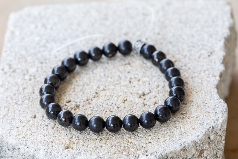 May include: A black beaded bracelet with a silver bead accent. The bracelet is made of round, smooth beads and is strung on a thin, clear cord.