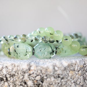 May include: A close-up of a green gemstone bracelet with black inclusions. The bracelet is made of round and oval-shaped beads. The beads are arranged in a layered pattern.