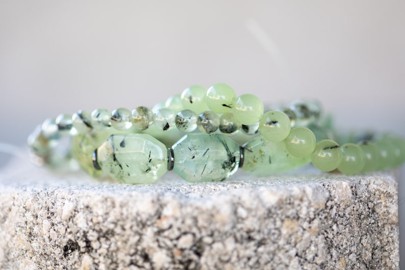 May include: A close-up of a green gemstone bracelet with black inclusions. The bracelet is made of round and oval-shaped beads. The beads are arranged in a layered pattern.