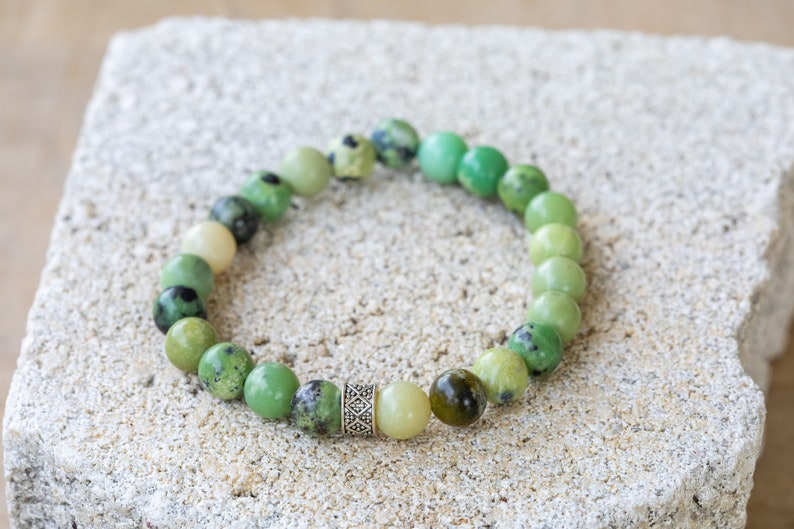 K&ouml;nnte beinhalten: Ein gr&uuml;nes Edelstein-Armband mit einem silbernen Anh&auml;nger. Das Armband besteht aus runden Perlen, die in der Gr&ouml;&szlig;e abgestuft sind, wobei die gr&ouml;&szlig;ten Perlen in der Mitte des Armbands liegen. Die Perlen sind in verschiedenen Gr&uuml;nt&ouml;nen gehalten, einige haben eine gesprenkelte Optik.