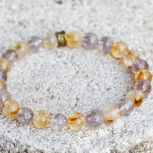 May include: A bracelet made of small, round, light purple and yellow beads. The beads are strung together on a clear elastic cord. The bracelet has a gold clasp.