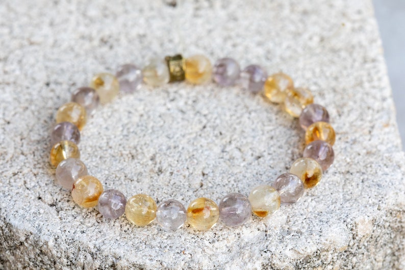 May include: A bracelet made of small, round, light purple and yellow beads. The beads are strung together on a clear elastic cord. The bracelet has a gold clasp.