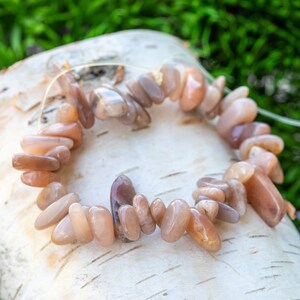 May include: A bracelet made of polished, peach-colored gemstone beads. The beads are strung together on a thin, clear cord. The bracelet is resting on a piece of birch bark, with green foliage in the background. The stones have a smooth, irregular shape.