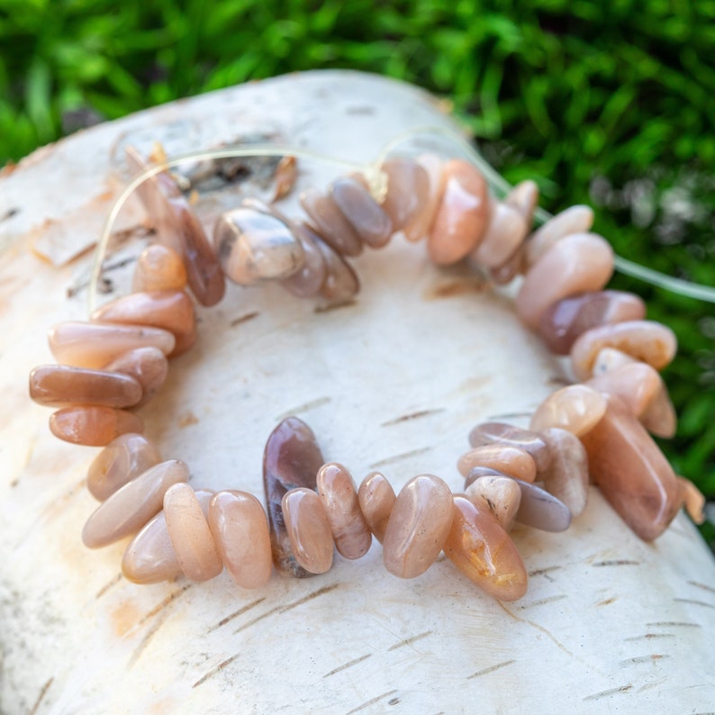 May include: A bracelet made of polished, peach-colored gemstone beads. The beads are strung together on a thin, clear cord. The bracelet is resting on a piece of birch bark, with green foliage in the background. The stones have a smooth, irregular shape.