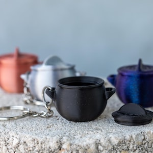 May include: A collection of miniature cauldron keychains in various colors, including copper, silver, black, and a blue-purple iridescent hue. Each cauldron has a small handle and is attached to a keychain ring. One cauldron has a detached lid. The keychains are displayed on a textured, light-colored surface.
