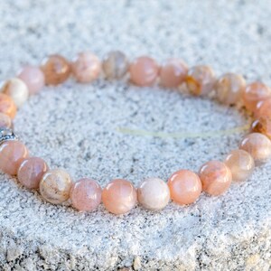 May include: A bracelet made of peach-colored gemstone beads. The beads are round and have variations in color, ranging from light peach to a darker orange. A small silver-colored spacer bead is visible.
