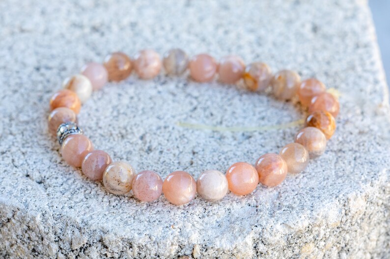 May include: A bracelet made of peach-colored gemstone beads. The beads are round and have variations in color, ranging from light peach to a darker orange. A small silver-colored spacer bead is visible.