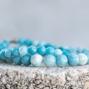 Peut inclure: Un bracelet en pierres bleues avec des perles rondes. Le bracelet est fait de pierres d'aigue-marine et repose sur une surface gris clair.