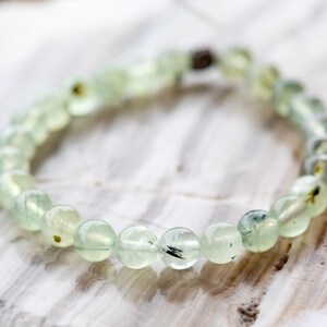 May include: A green gemstone bracelet with a silver clasp. The bracelet is made of round beads with black and green inclusions.