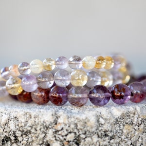 May include: Three beaded bracelets made of amethyst, citrine, and clear quartz gemstones. The bracelets are stacked on top of each other on a grey stone surface.
