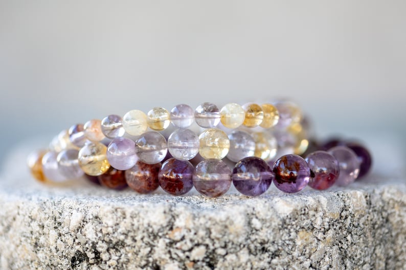 May include: Three beaded bracelets made of amethyst, citrine, and clear quartz gemstones. The bracelets are stacked on top of each other on a grey stone surface.