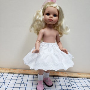 May include: A white doll with blonde hair and blue eyes wearing a white lace petticoat and pink shoes. The doll is standing on a white and black checkered surface.