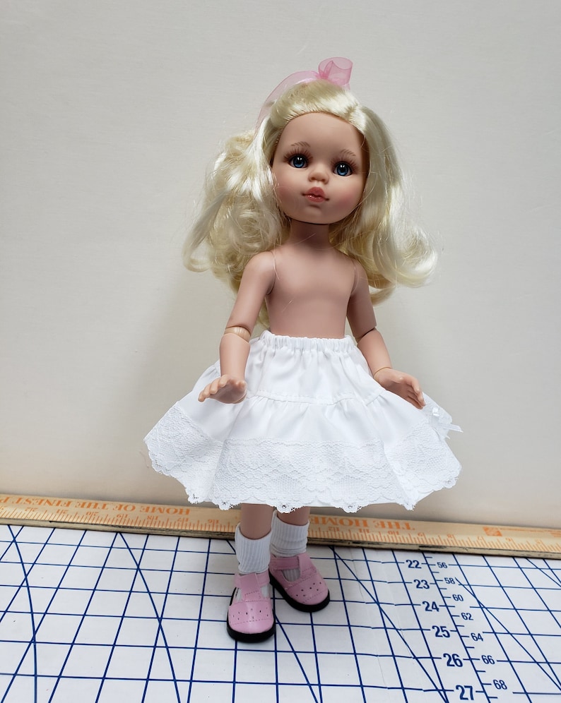 May include: A white doll with blonde hair and blue eyes wearing a white lace petticoat and pink shoes. The doll is standing on a white and black checkered surface.