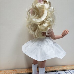 May include: A white doll with blonde curly hair wearing a white skirt and pink shoes. The doll is standing on a checkered surface.