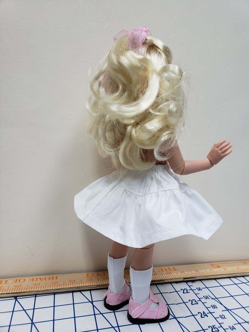 May include: A white doll with blonde curly hair wearing a white skirt and pink shoes. The doll is standing on a checkered surface.