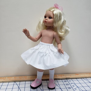 May include: A white doll skirt with a ruffled hem and a small bow at the waist. The skirt is worn by a doll with blonde hair and blue eyes. The doll is wearing pink socks and shoes.