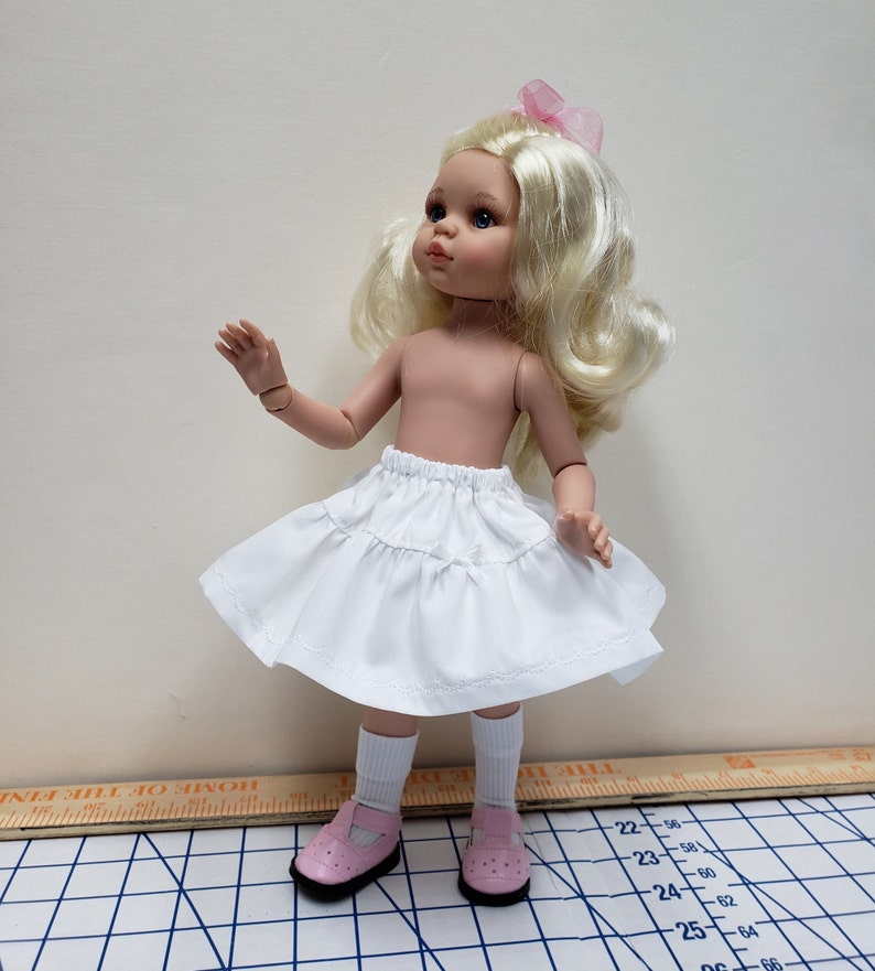 May include: A white doll skirt with a ruffled hem and a small bow at the waist. The skirt is worn by a doll with blonde hair and blue eyes. The doll is wearing pink socks and shoes.