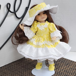 May include: A doll wearing a yellow dress with white floral accents and a matching sun hat. The dress has a white lace trim and the doll is wearing yellow shoes. The doll has long brown hair and is standing on a white stand.
