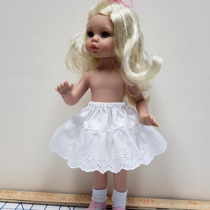 May include: A white doll with blonde hair wearing a white lace skirt and pink shoes. The doll is standing on a checkered surface.