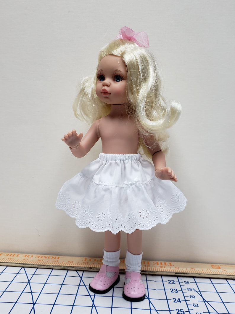 May include: A white doll with blonde hair wearing a white lace skirt and pink shoes. The doll is standing on a checkered surface.